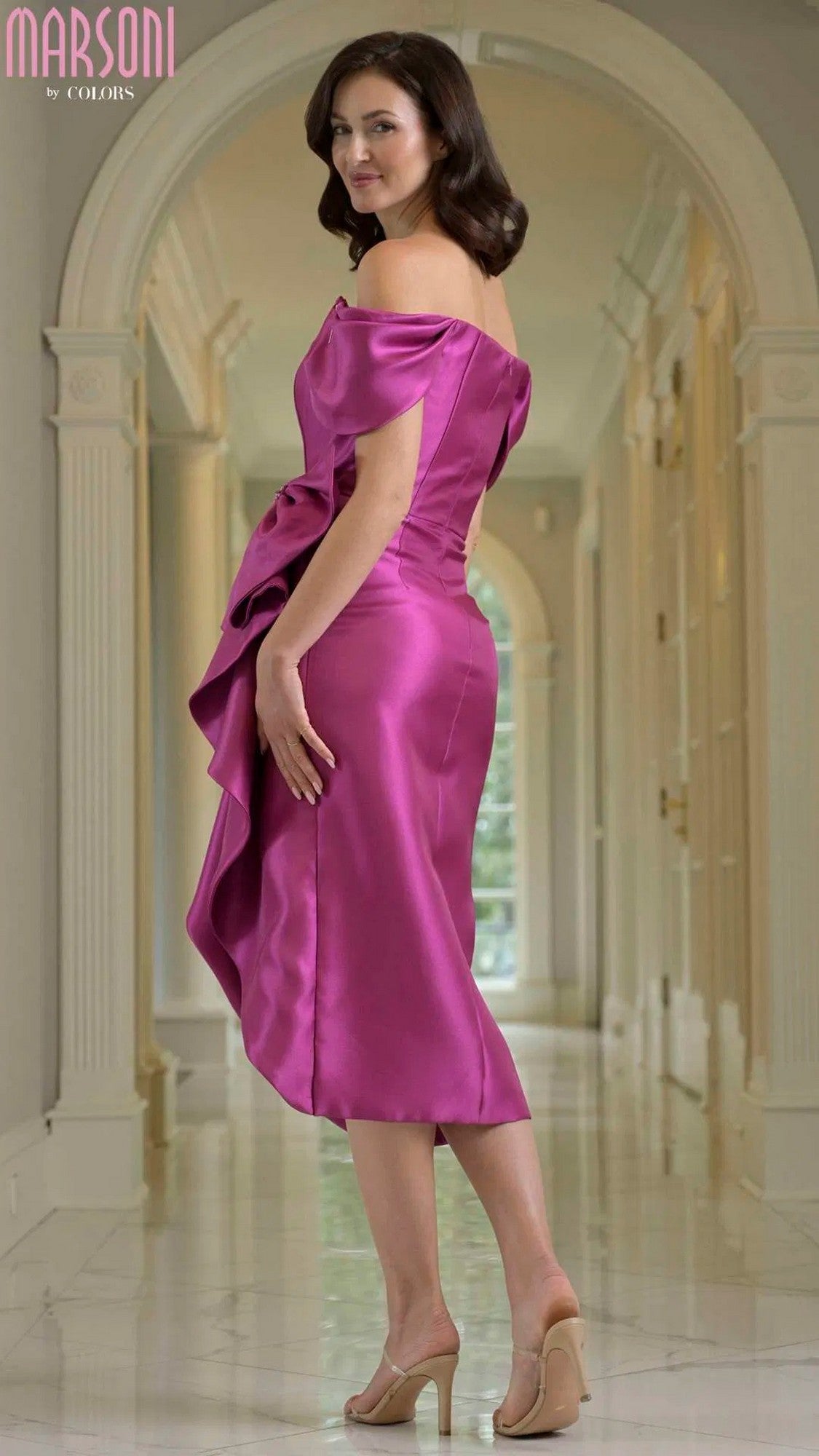A woman wearing the Marsoni MV1389 knee-length semi-formal fuchsia dress with off-shoulder sleeves and beige heels stands in an elegant hallway with arches and columns, glancing over her shoulder.